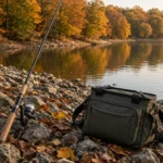 A spinning rod with a spinnerbait and a small tackle bag on a rocky bank beside a lake in autumn