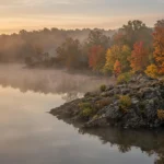 Mist rising off a calm reservoir lined with autumn trees on a cool fall morning