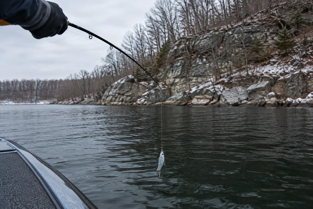 A gloved hand lifts a rod with a silver blade bait over deep water beside a snowy rocky bluff in winter.