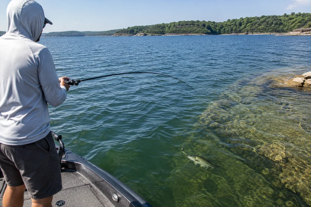 A baitcasting rod sweeps a bottom rig along a steep gravel drop-off on a reservoir in bright midday sun.