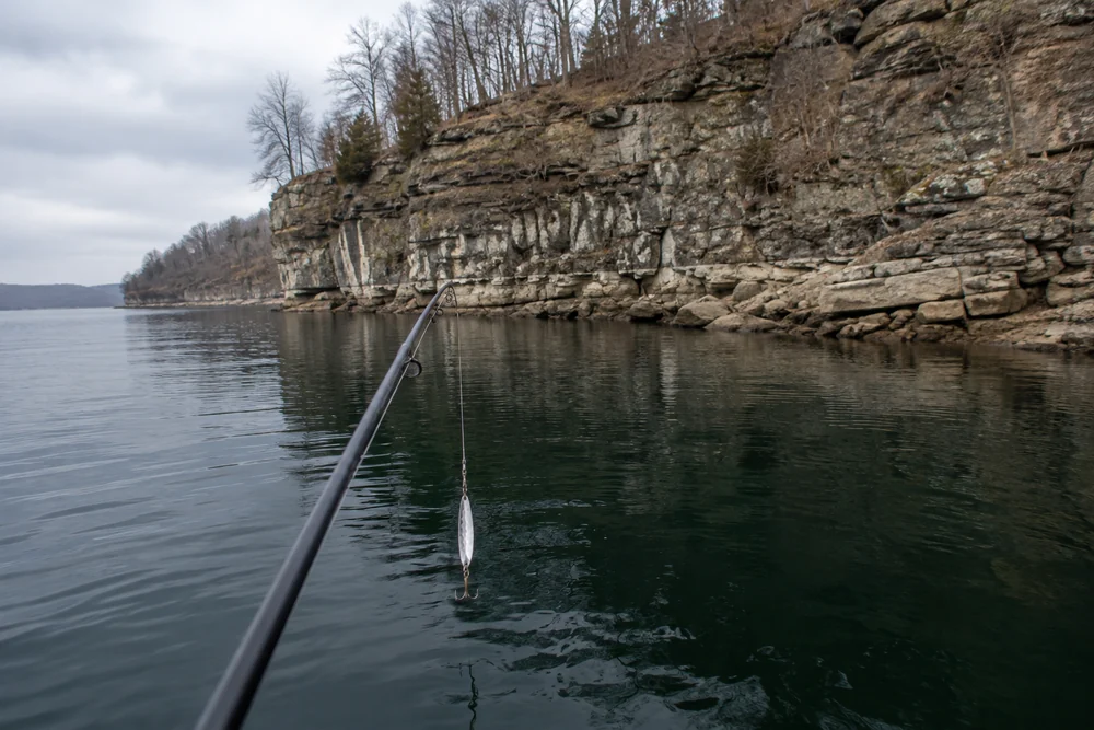 A rod tip holding a silver jigging spoon above deep water beside a rocky bluff on an overcast winter reservoir.